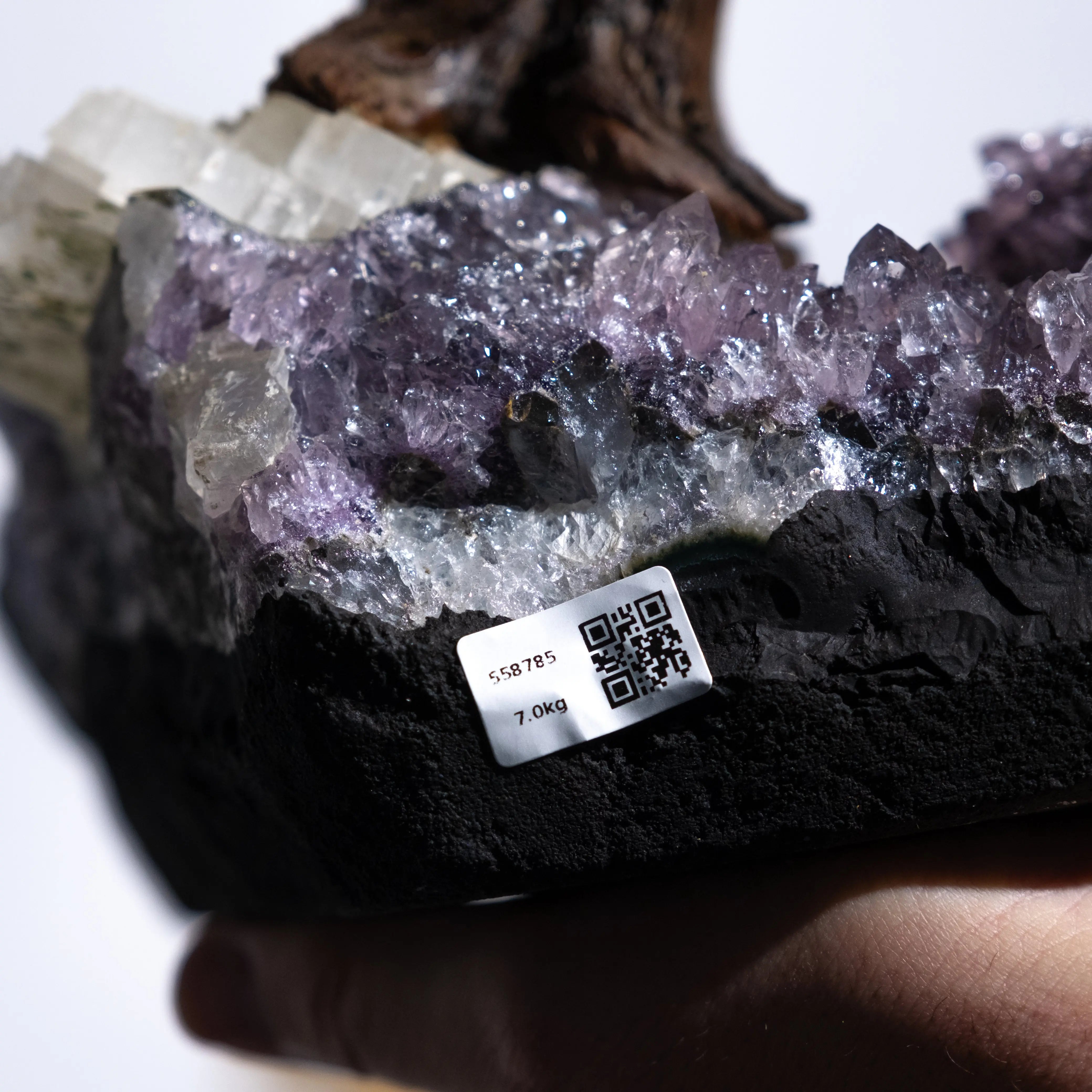 Close-up of a hand holding a crystal cluster with a QR code and weight label, showing purple and white shimmering crystals.
