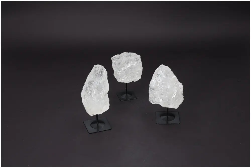 Three rough clear quartz crystals on metal stands, used for purification and harmonizing, displayed on a dark background.