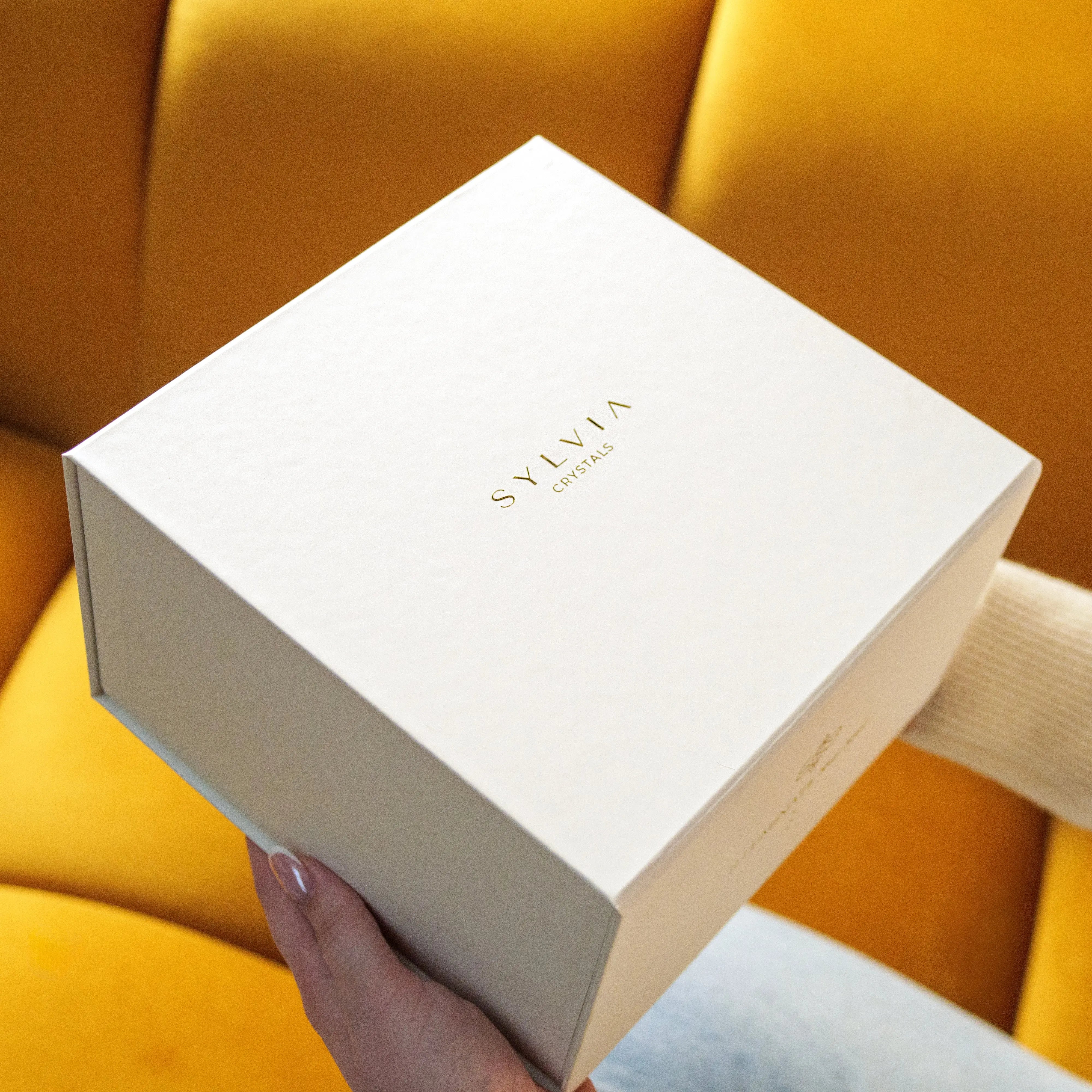 Elegant beige gift box labeled 'Sylwia' on a yellow couch, part of the Now and Forever Gift Box set with crystals and a luxury candle.