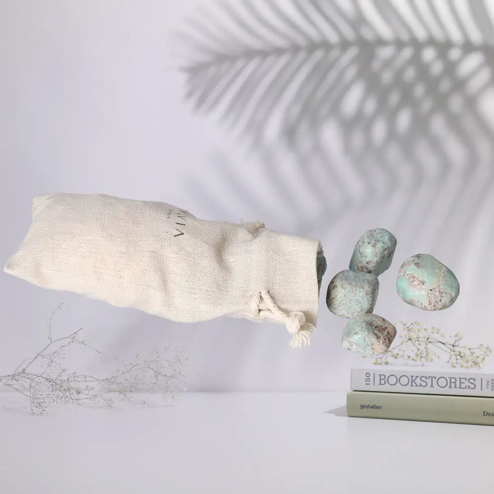 Tumbled Amazonite stones emerging from a cloth bag, showcasing their unique turquoise and green patterns, placed beside a book.