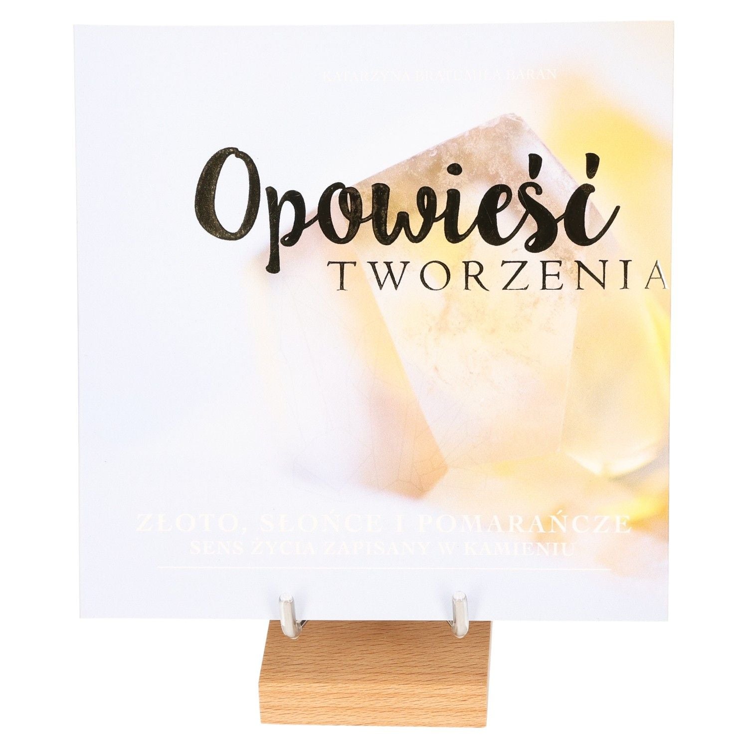 Book cover "Opowieść Tworzenia" with abstract sunlight and stone design on a wooden stand.